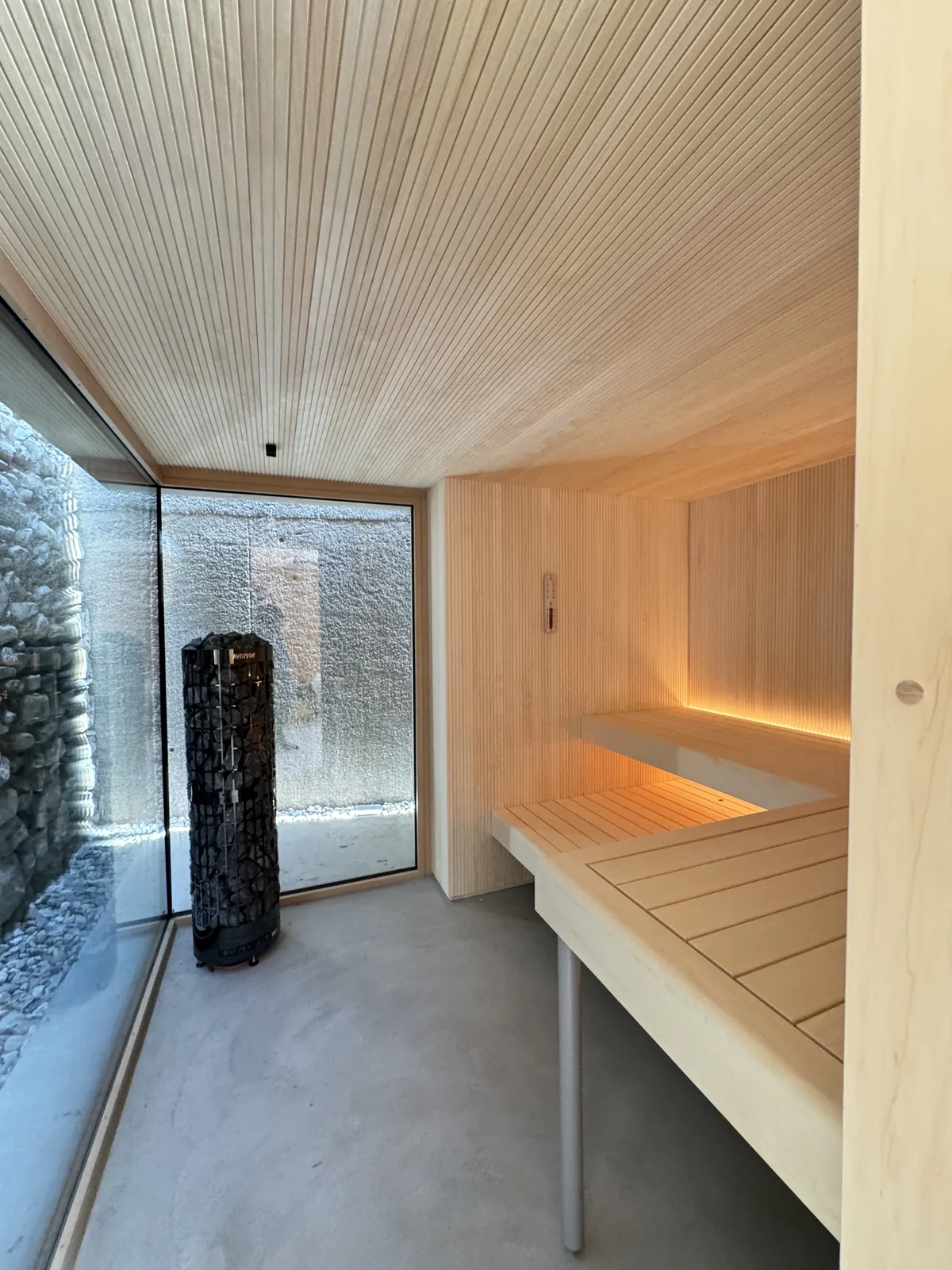 Helle Sauna mit Panorama-Glasfront, Säulenofen und beleuchteten Liegebänken
